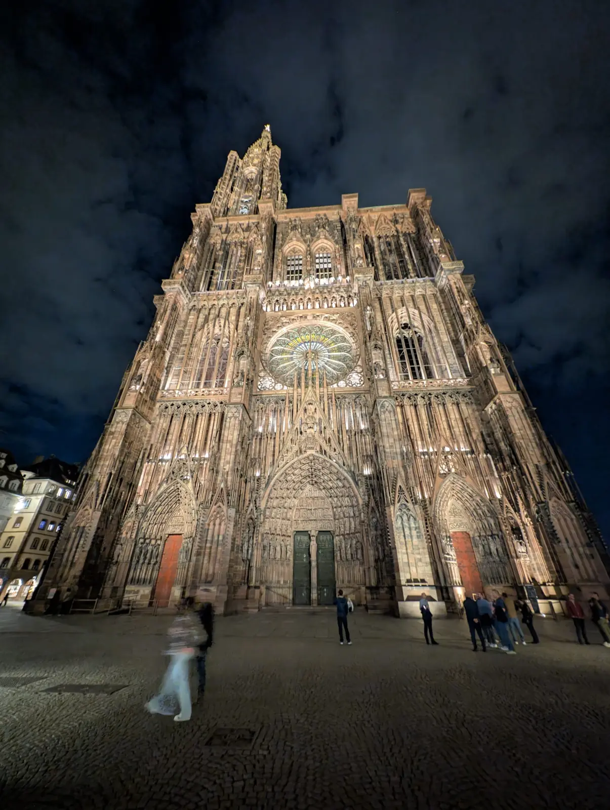 The floodlit west façade and spire of Strasbourg Cathedral at night, with walkers blurred into ghosts in the square