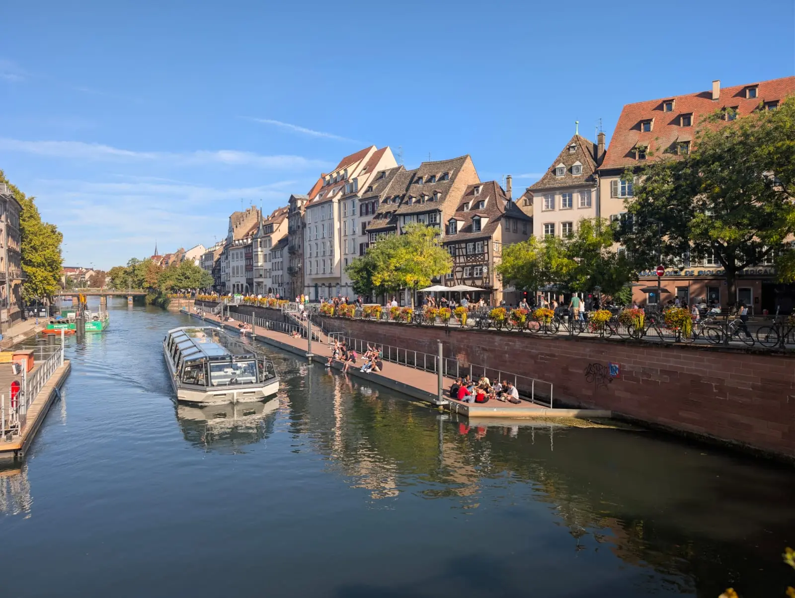 A long view down the river Ill with a glass-roofed tour boat, a floating pontoon and tall houses lining the quay