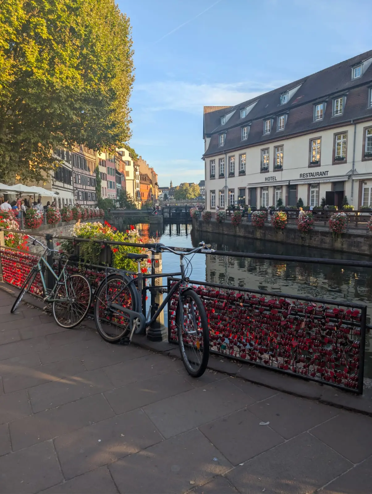 Two bicycles leaning on a canal railing covered in red heart-shaped padlocks, with the canal leading away between buildings